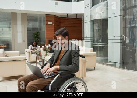 Young disabled businessman sitting in wheelchair and working online using laptop Stock Photo