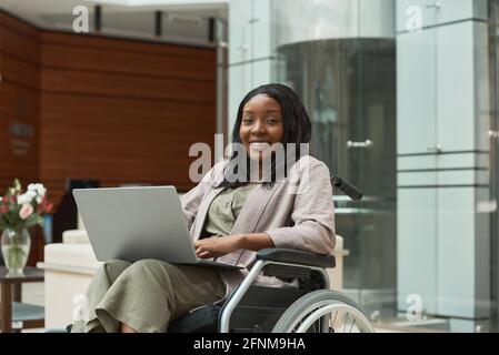 Portrait of African disabled woman smiling at camera while sitting in wheelchair and working on laptop Stock Photo