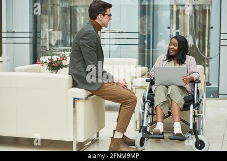 Businessman talking to his colleague while she sitting in wheelchair and working on laptop at office Stock Photo