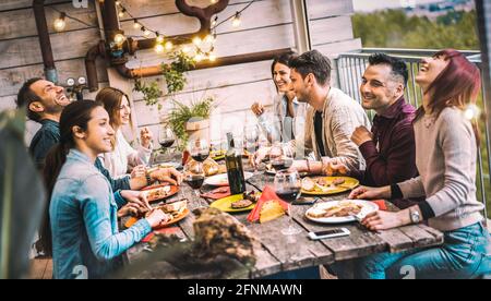 Young people dining and having fun drinking red wine together on balcony rooftop dinner party - Happy friends eating bbq food at restaurant patio Stock Photo