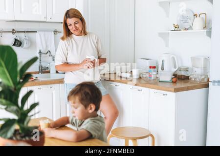 Young woman mother looking as her toddler boy son having breakfast at the table in bright kitchen at home Stock Photo