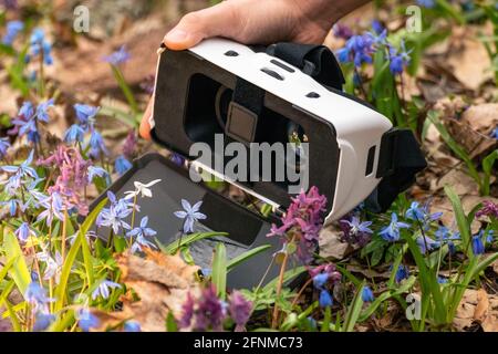 Hand holding 3D Virtual reality headset glasses with lenses opened in spring blue and purple wild flowers. VR tech gear goggles close-up. Optical syst Stock Photo