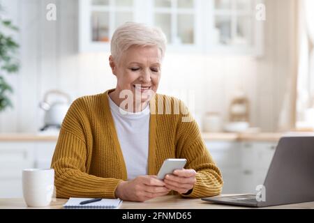 Smiling mature lady sitting at table with laptop, using smartphone Stock Photo