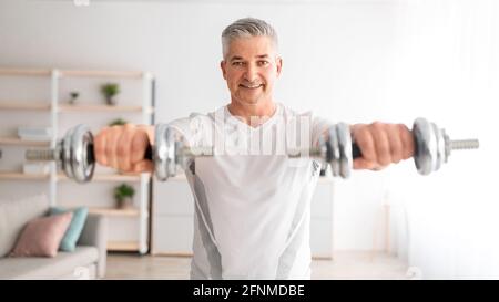 Athletic mature man lifting dumbells up, doing workout on arms strength, training at home in living room interior Stock Photo