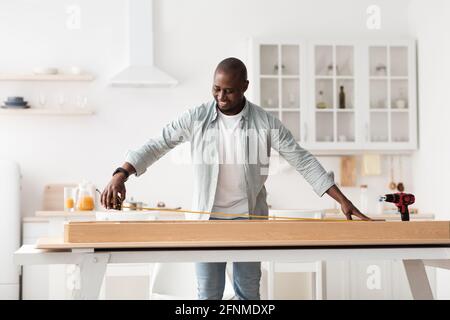 Assembling furniture. Happy african american man measuring table with roulette, standing and installing desk in kitchen Stock Photo