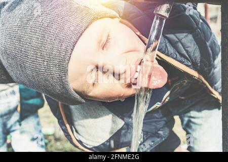 Funny little boy in grey knitted hat drinks water from vintage water pump in village house yard on sunny day close view Stock Photo