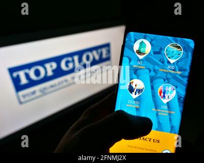 Person holding smartphone with webpage of Malaysian company Top Glove Corporation Berhad on screen in front of logo. Focus on center of phone display. Stock Photo