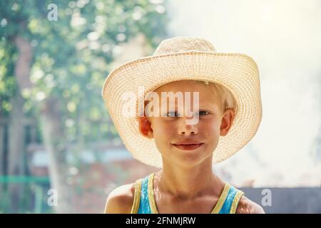 Cheerful little boy wearing large straw hat and blue sleeveless shirt posing for camera in sunny summer city park close view Stock Photo
