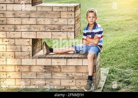 Positive little boy in striped polo t-shirt and denim shorts sits on decorative wooden installation on lush green meadow in park Stock Photo