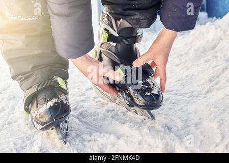 Boy in warm jacket and pants adjusts ice skates standing on snowy ground near rink on cold day extreme close view Stock Photo