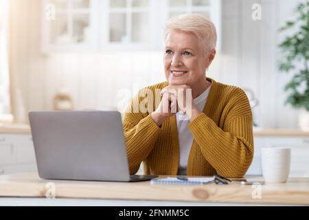 Happy old lady working on laptop at home, kitchen interior Stock Photo