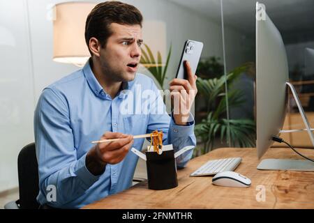Confused handsome young business man using mobile phone while having lunch at the work Stock Photo