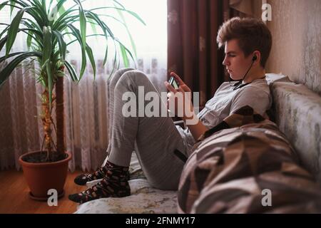 A young guy, a teenager sit on the sofa at home in headphone and writes sms on a social network or listen music on a smartphone. Stock Photo