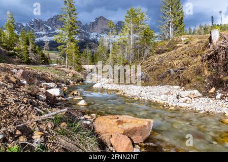 Mountain creek with snowy rocky peaks in background under stormy sky in spring. A Wooden footbridge over the creek is visible in distance. Stock Photo