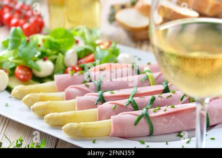 Delicious spring salad with asparagus rolled in cooked ham on a white platter, served with a young dry white wine and baguette slices Stock Photo