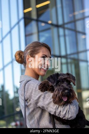 Pretty young business woman holding cute dog in arms in front of office building Stock Photo