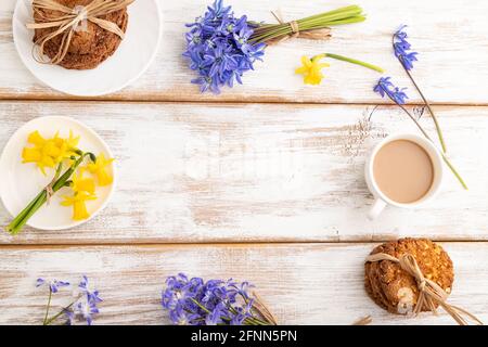 Frame composition with oatmeal cookies, spring snowdrop flowers bluebells, narcissus and cup of coffee on white wooden background. top view, flat lay, Stock Photo