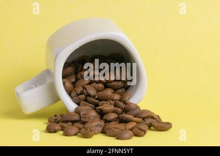 White overturned cup of coffee with roasted coffee beans inside on yellow background Stock Photo