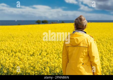 rear view of woman in yellow raincoat against canola field and sea on sunny day Stock Photo