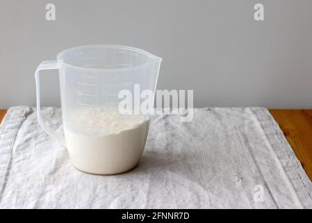 Plastic measuring cup with flour stands on a linen tablecloth on a wooden table against a gray blurred wall. Selective focus Stock Photo