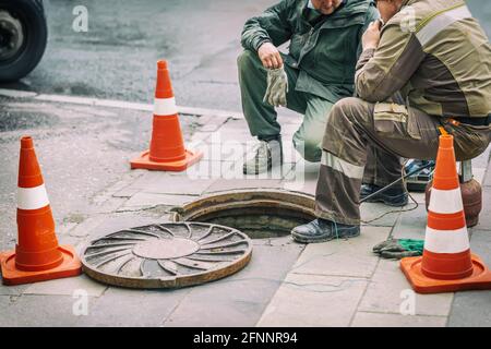 Workers sitting on over open sewer hatch on street near traffic cones. Repair of sewage, underground utilities concept Stock Photo