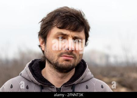 A handsome bearded man with disheveled hair squinted his eye in displeasure against a blurred background. Man in a jacket with a hood Stock Photo