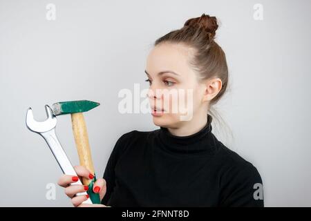 A woman with bunched hair and a black sweater looks at a hammer and a big wrench, while hoding them in her hands Stock Photo