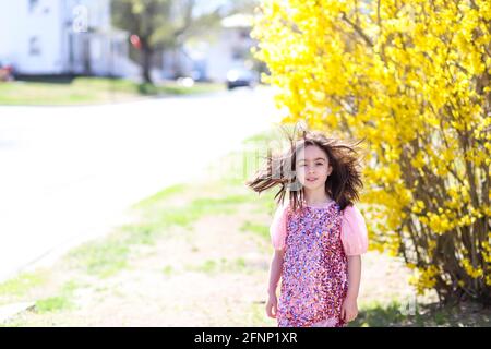 Girl in a sparkly dress stands in front of a forsythia on a windy day