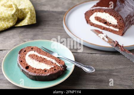 A slice of Gluten Free Chocolate Swiss roll with a mascarpone whipped cream filling and chocolate ganache topping, plated in front of the main cake Stock Photo