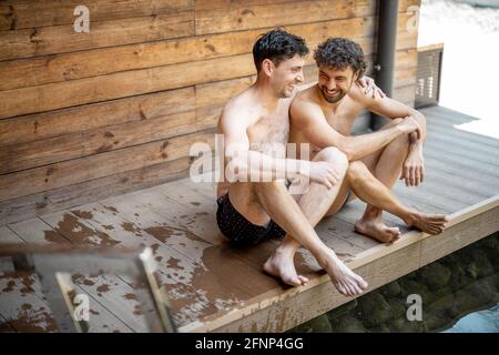 Male friends sitting on the terrace near the lake in the spa complex and talking. Relax and fun time in wellness spa. Hugs and laughing together. Stock Photo