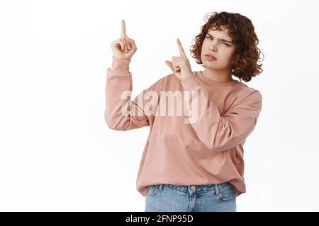Confused girl points at upper left corner, looks puzzled and clueless, need answers, asking question about advertisement on top, what is this Stock Photo