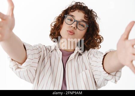 Close up portrait of cute young woman in glasses stretch out hands, holding camera as if taking selfie or video chat with someone, making kissing lips Stock Photo