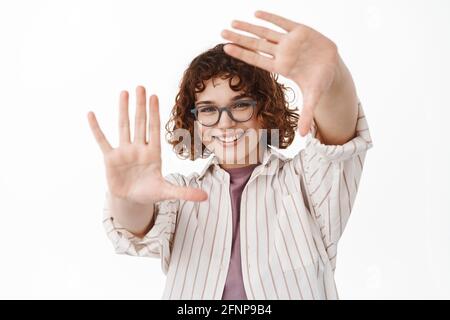 Lets get creative. Smiling young woman imaging something, wearing glasses, looking through hand frames camera gesture, searching for perfect shot Stock Photo