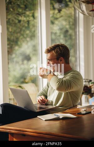 Man having coffee while working from home. Man sitting at table with laptop drinking coffee looking away and smiling. Stock Photo