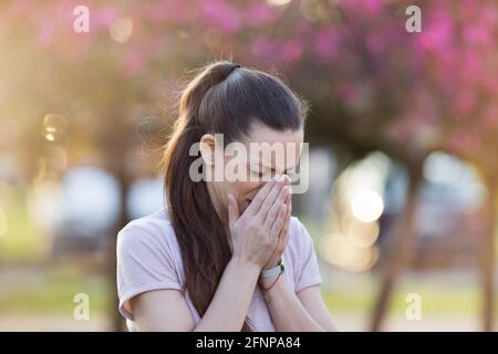 Young pretty woman sneezing in front of blooming tree. Spring allergy concept Stock Photo