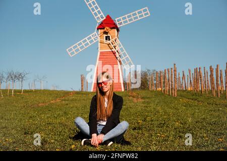 Beautiful young woman in red sunglasses sits on the grass against the backdrop of the red mill. Sunny summer day. Rest in the countryside Stock Photo