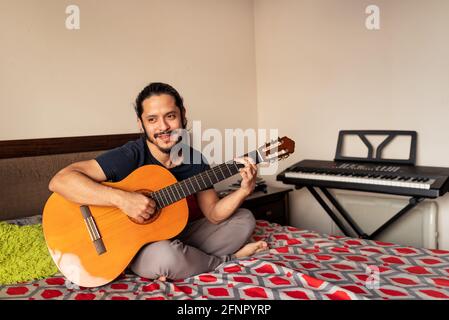Young latin man sitting on bed playing acoustic guitar at home in the bedroom Stock Photo