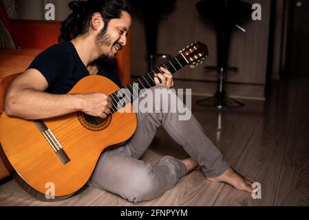 Young latin man sitting on the floor playing guitar at home in the living room Stock Photo