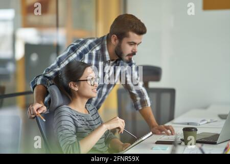 Professional male mentor or teacher explaining computer work to young asian female intern while working together in the modern coworking space Stock Photo