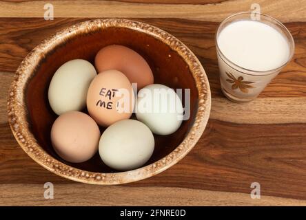 A brown bowl of eggs with a small glass of milk.  A humorous phrase is written on one of he eggs. Stock Photo