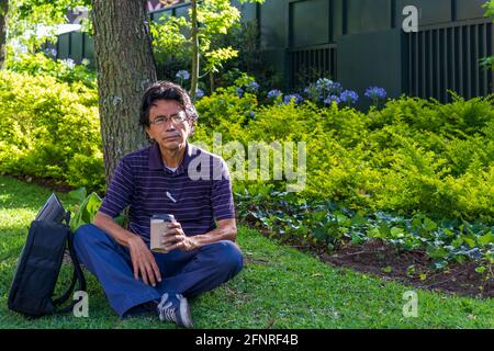 Hispanic man wearing a face mask on a park drinking coffee Stock Photo