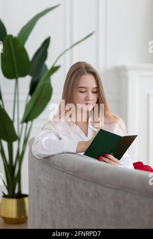 Blonde long haired young Caucasian business woman writing in planner notepad, diary, planning work and affairs schedule, sitting on sofa, vertical sho Stock Photo
