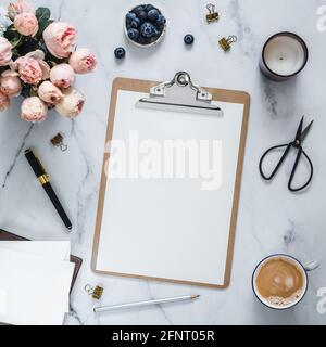 Top view of clipboard with white empty page. Clipboard, flowers, scented candle on white marble. Feminine home office mockup with blank sheet of paper A4 portrait format, copy space. Flat lay Stock Photo