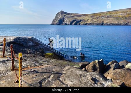 Three scuba divers climbing up steps in Port Erin Isle of Man with Bradda Head in the background Stock Photo