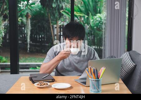 Young asian man holding a cup of coffee and using laptop while sitting near window in home office. Stock Photo