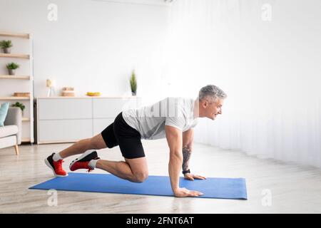 Stay fit. Athletic senior man training and doing running plank exercise on mat in living room at home, empty space Stock Photo