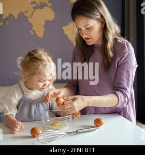 Involving children in cooking process. Young mom showing cute little daughter how to break fresh eggs into bowl for pie or omelet while preparing food together at home. Mother teaching kid to cook Stock Photo