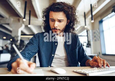 Handsome curly guy is making notes at the working table in open space office Stock Photo