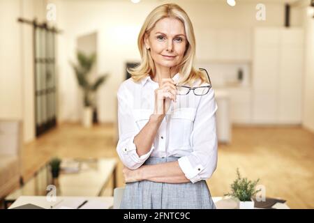 Portrait of confident mature blonde businesswoman looking at camera, holding glasses while posing, standing in the office Stock Photo