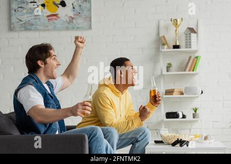 KYIV, UKRAINE - MARCH 22, 2021: interracial friends sitting on couch with bottles of beer and emotionally watching tv in living room Stock Photo